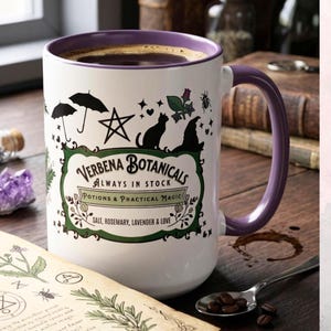 Könnte beinhalten: Ein weißer Keramikbecher mit violettem Innenraum und Henkel, verziert mit schwarzen Illustrationen von Regenschirmen, einem Stern, einer Katze und einem Hexenhut. Der Text lautet Verbena Botanicals, Always in Stock, Potions & Practical Magic.