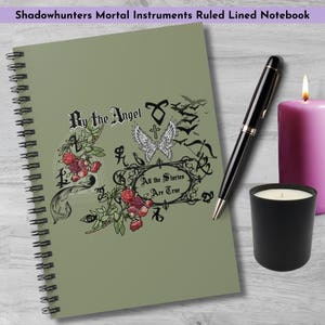 May include: A sage green spiral-bound notebook with the text "By the Angel" and "All the Stories Are True." A black pen, a black candle, and a pink candle are next to the notebook. The notebook is labeled "Shadowhunters Mortal Instruments Ruled Lined Notebook."