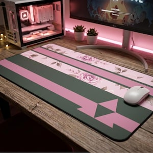 Peut inclure: Un tapis de bureau rectangulaire avec une base gris foncé, des bandes horizontales rose clair et un motif floral de roses roses et de feuilles vertes. Une souris d'ordinateur blanche repose sur le côté droit du tapis, orné d'un motif triangulaire.