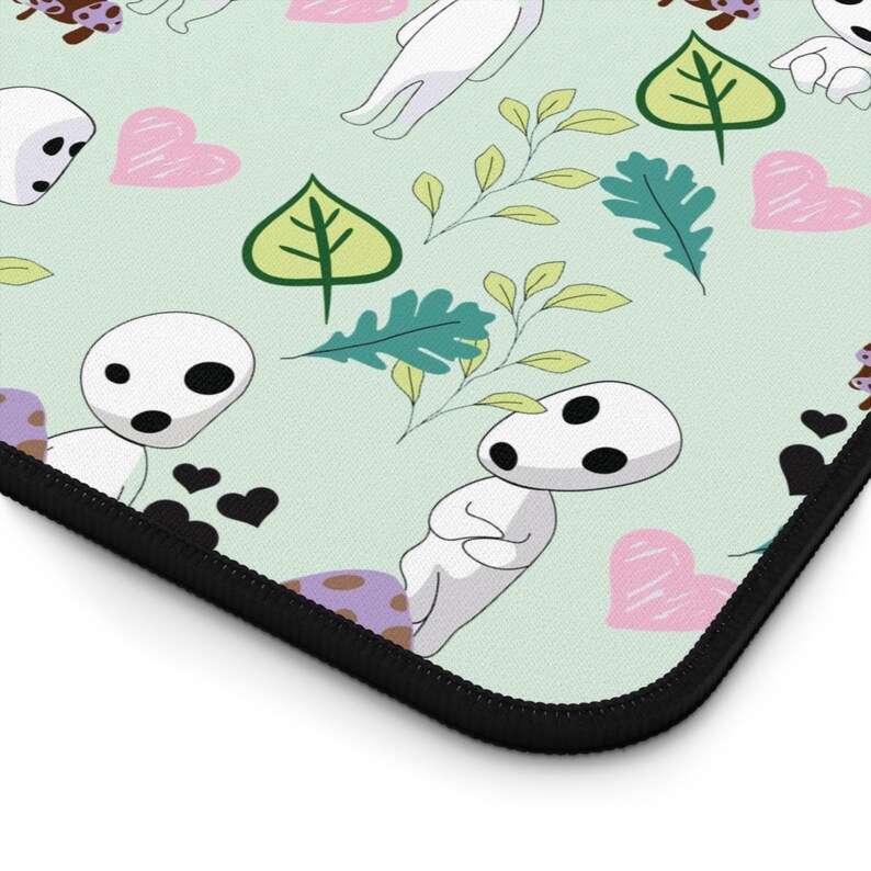 May include: A light green mousepad featuring a repeating pattern of white spirit-like figures with black eyes, pink hearts, green leaves, and sprigs of foliage. The mousepad has a black stitched edge.