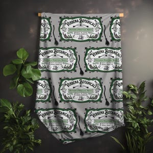 May include: A gray fabric with a repeating pattern of vintage-style labels and broom illustrations. The labels read "VERBENA BOTANICALS" with text about potions and practical magic. The fabric is draped over a wooden rod.
