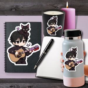 May include: A black spiral notebook, a black candle, a black pen, and a light gray water bottle, all featuring a sticker of a black-haired character playing an acoustic guitar with a black cat on their head and the text MIN AA & TANG.