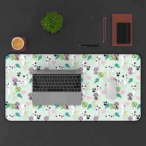 May include: A light green desk mat featuring a repeating pattern of white spirit-like figures, purple mushrooms, pink hearts, and green leaves. A laptop, mouse, and other office supplies are on the desk.