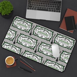 May include: A gray mousepad featuring a repeating pattern of green and white labels with the text "VERBENA BOTANICALS" and small black witch brooms. A computer mouse, keyboard, and other office supplies are visible.
