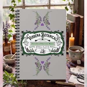 May include: A gray spiral-bound notebook featuring a central green and black label with the text Verbena Botanicals, Potions & Practical Magic, Coupeville, Always in Stock, Massachusetts, Salt, Rosemary, Lavender & Love, surrounded by lavender.