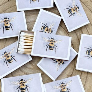 May include: A collection of matchboxes with a watercolor bee design. The boxes are white with a light gray background and a detailed illustration of a bee. One box is open, revealing the wooden matchsticks.