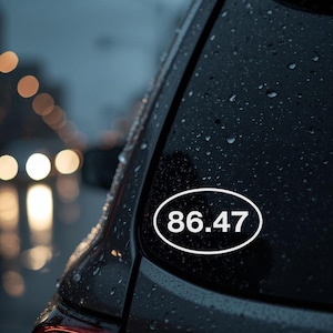 May include: An oval white sticker with the number 86.47 in black is affixed to the rear window of a dark-colored car. The background shows a blurred street scene with lights and rain.