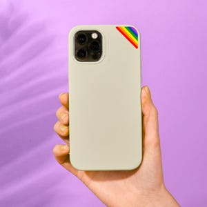 May include: A light beige smartphone case featuring a triple-lens camera cutout on the upper left and a small, diagonal rainbow-colored stripe accent located in the top right corner. The case has a smooth, matte finish and a rectangular shape.