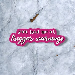 May include: Pink sticker with white lettering on a gray marble background. The sticker says "You had me at trigger warnings."
