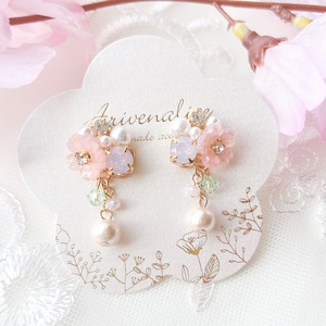 Peut inclure: Une paire de boucles d'oreilles fleuries. Chaque boucle d'oreille est ornée de fleurs roses, de cristaux transparents et de perles blanches pendantes. Les boucles d'oreilles sont présentées sur une carte blanche avec du texte doré et des motifs floraux.