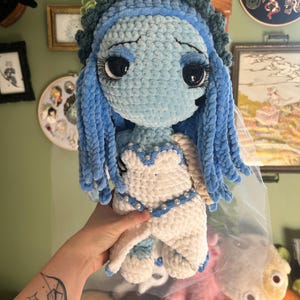 May include: A handmade crochet doll in the style of a character from a stop-motion animated film. The doll features a light blue face and hair, a white dress with pearl accents, and large black eyes. The doll is approximately 10 inches tall.
