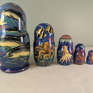 May include: A set of six nesting dolls, each painted with marine life scenes. The largest doll features dolphins leaping from the water, while others depict an octopus, fish, and seahorses. The dolls are primarily blue with colorful accents.