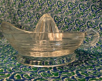 May include: A clear glass citrus juicer with a ribbed design and a handle on one side. The juicer has a central cone for squeezing citrus fruits.