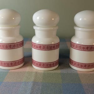 May include: Three white ceramic jars with red and white patterned bands. The jars have a rounded top and a wide mouth.