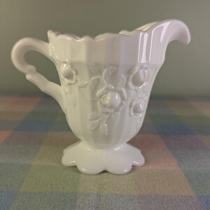 May include: A white milk glass creamer with a handle and a spout. The creamer features embossed floral designs and a scalloped rim. The creamer sits on a pedestal base with a scalloped edge. The creamer is likely vintage and is a decorative item.