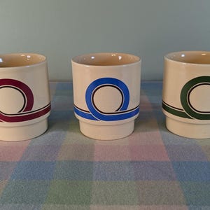 May include: Three cream-colored ceramic mugs with a retro design. Each mug features a different colored loop design: burgundy, blue, and green. The mugs have a stepped base and a simple, cylindrical shape.