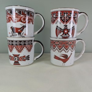 May include: Four white ceramic mugs with brown and red Southwestern-style designs. Each mug features a different animal or geometric pattern, with a speckled finish and dark brown rim and handle. The mugs are stacked in pairs.
