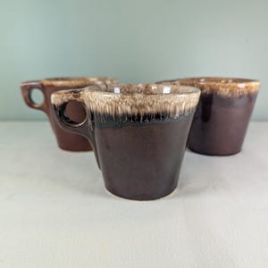 May include: Three brown ceramic mugs with a unique design. The mugs have a dark brown base that fades to a lighter brown at the rim. Each mug has a small handle. The mugs are arranged on a white surface.