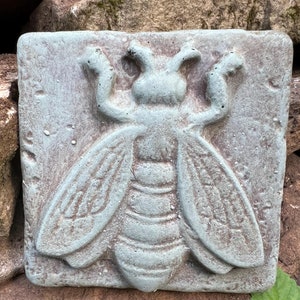 May include: A square, light blue, concrete garden tile with a raised, detailed design of a bee. The bee has a textured body and wings.