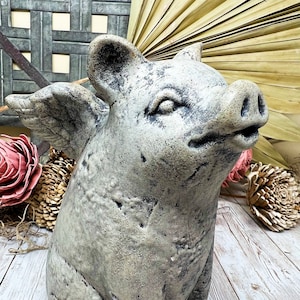 May include: A gray concrete pig figurine with wings. The pig is sitting on a white wooden surface. The pig has a textured surface and a realistic appearance.