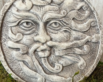 May include: A white, circular, concrete sculpture of a face with a stylized, swirling design. The face has a large nose, a wide mouth, and two eyes. The sculpture is set against a background of green moss.