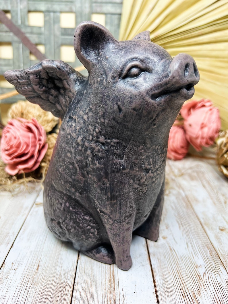May include: A gray ceramic figurine of a pig with wings. The pig is sitting with its wings spread out. The figurine has a textured surface and a realistic appearance.