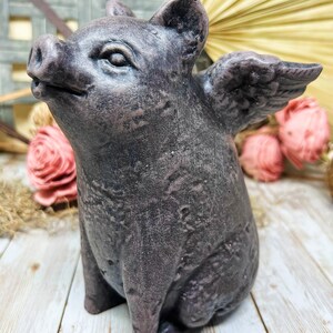 May include: A small, gray, winged pig figurine with a textured surface. The pig is sitting on a white wooden surface.