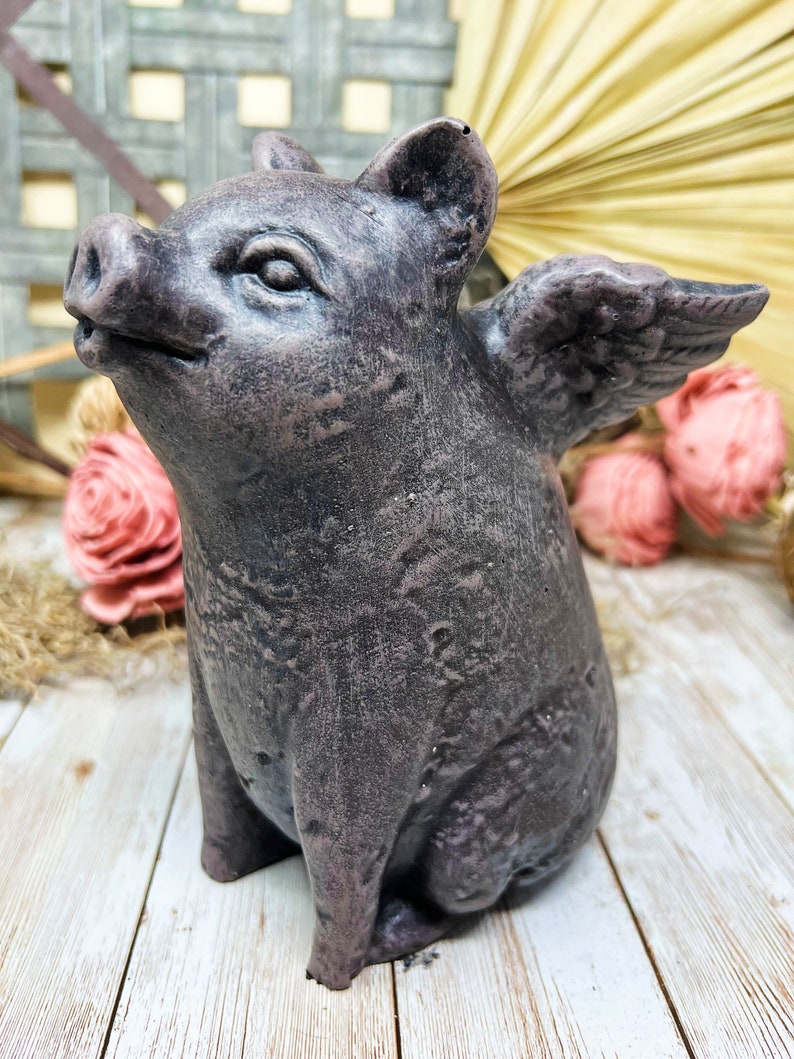 May include: A small, gray, winged pig figurine with a textured surface. The pig is sitting on a white wooden surface.