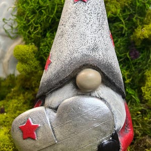 May include: A whimsical garden gnome statue holding a heart. The gnome wears a gray hat with red stars and a red coat. The heart is silver with a wood grain texture. The gnome's nose is beige, and the base is red.