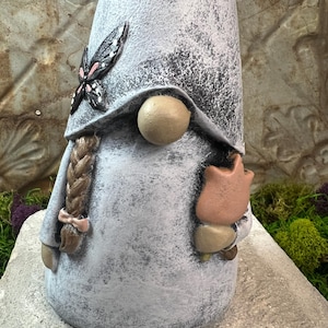 May include: A white gnome with a gray and white speckled finish. The gnome is wearing a pointed hat with a black butterfly design and holding a pink flower.