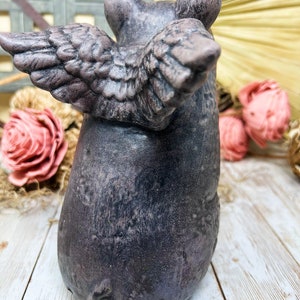 May include: A gray ceramic pig figurine with wings. The pig is standing on a white wooden surface. The pig has a textured surface and a detailed face.