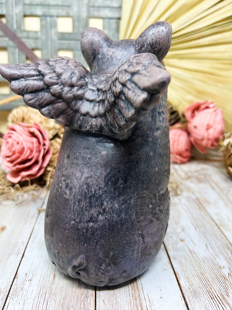 May include: A gray ceramic pig figurine with wings. The pig is standing on a white wooden surface. The pig has a textured surface and a detailed face.