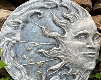 May include: A round, gray stone sculpture of the sun and moon with a blue background and gold stars. The sun has a human face and the moon is a crescent shape.