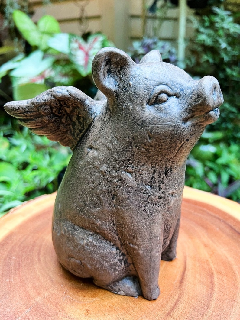 May include: A gray concrete figurine of a pig with wings. The pig is sitting with its head tilted up and its wings spread out behind it.