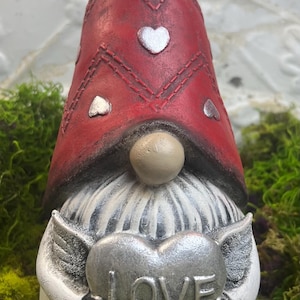May include: A Valentine's Day gnome figurine with a red hat adorned with silver hearts. The gnome holds a silver heart with the word "LOVE" on it. The figurine has a white beard and is set against a backdrop of green moss.