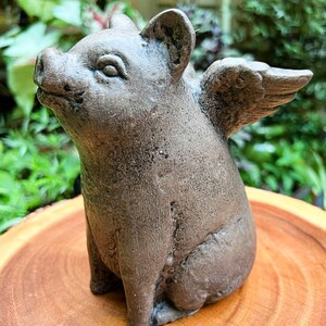 May include: A gray ceramic pig figurine with wings. The pig is sitting on a wooden surface.