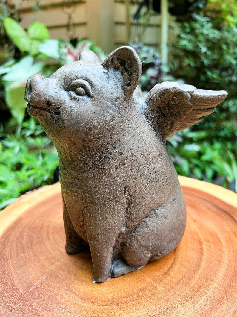 May include: A gray ceramic pig figurine with wings. The pig is sitting on a wooden surface.