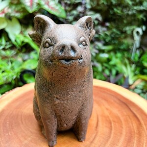 May include: A small, brown ceramic pig figurine with a friendly expression. The pig is sitting on a wooden surface.