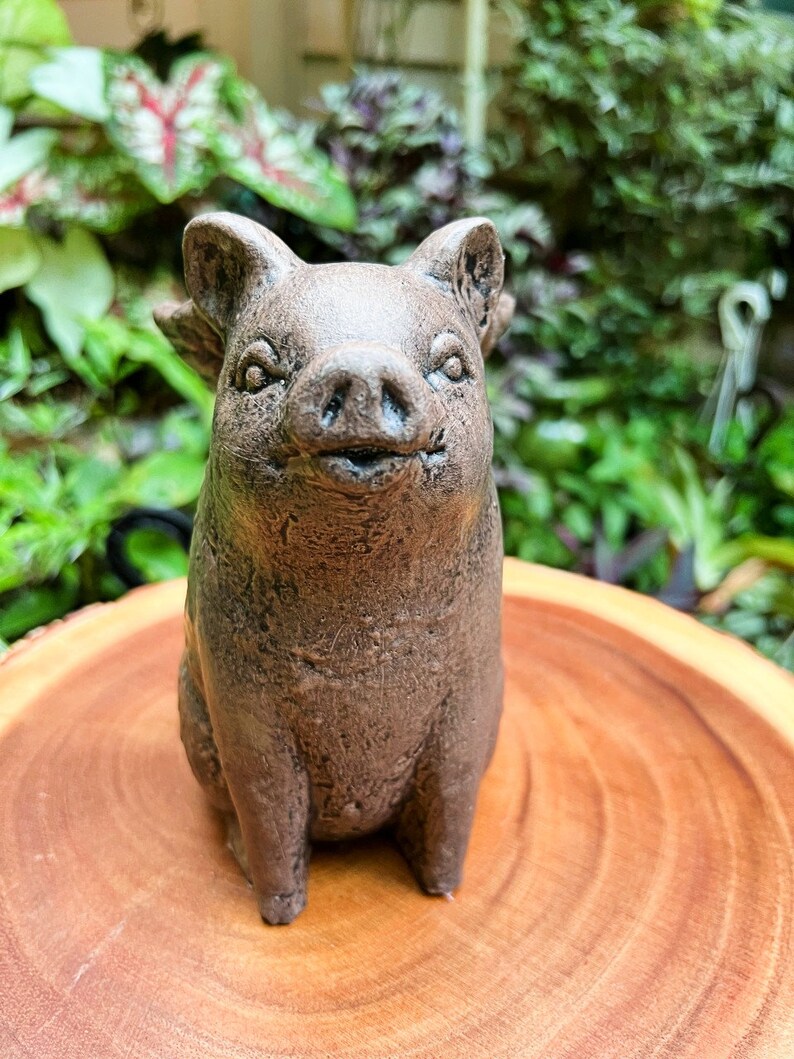 May include: A small, brown ceramic pig figurine with a friendly expression. The pig is sitting on a wooden surface.
