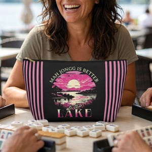 May include: A black and pink striped cosmetic bag with the text "Mah Jongg is Better at the Lake" and a sunset graphic. The bag is held by a person smiling while playing a game of Mahjongg.