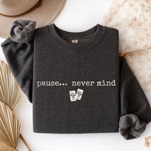 May include: A dark charcoal gray crewneck sweatshirt featuring the white text "pause... never mind" printed across the chest. Below the text are two small white rectangular graphics depicting tarot cards with a central design element.