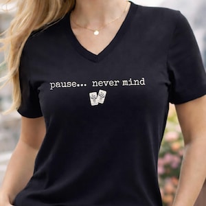 Pode incluir: Uma t-shirt preta com decote em V que apresenta o texto branco "pause... never mind" impresso no peito, acompanhado por um pequeno gr&aacute;fico de duas pe&ccedil;as retangulares brancas com um design de bambu centrado abaixo do texto.