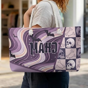 May include: A tote bag featuring a split design with purple and cream wavy lines, silhouettes of flying bats, and the text "MAHJ" in a dark font. The other side displays a checkered pattern of light pink squares and dark skulls with bones.