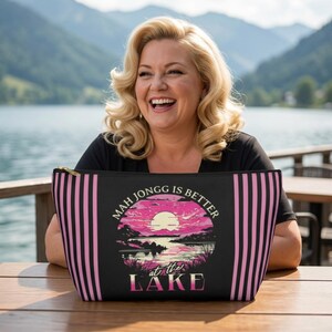 May include: Black zippered cosmetic bag with pink vertical stripes and a circular graphic. The graphic features a sunset over a lake with the text "MAH JONGG IS BETTER at the LAKE".