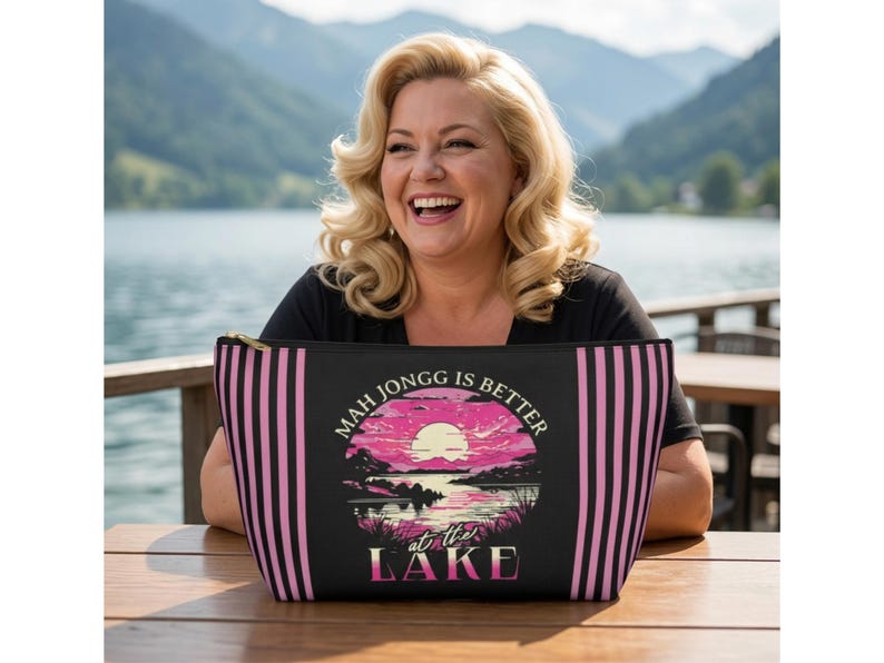 May include: Black zippered cosmetic bag with pink vertical stripes and a circular graphic. The graphic features a sunset over a lake with the text "MAH JONGG IS BETTER at the LAKE".