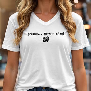 Pode incluir: Uma t-shirt branca com decote em V apresentando o texto preto "pause... never mind" impresso no peito. Abaixo do texto, h&aacute; dois pequenos &iacute;cones retangulares pretos, cada um exibindo um gr&aacute;fico branco de um cacto num vaso.