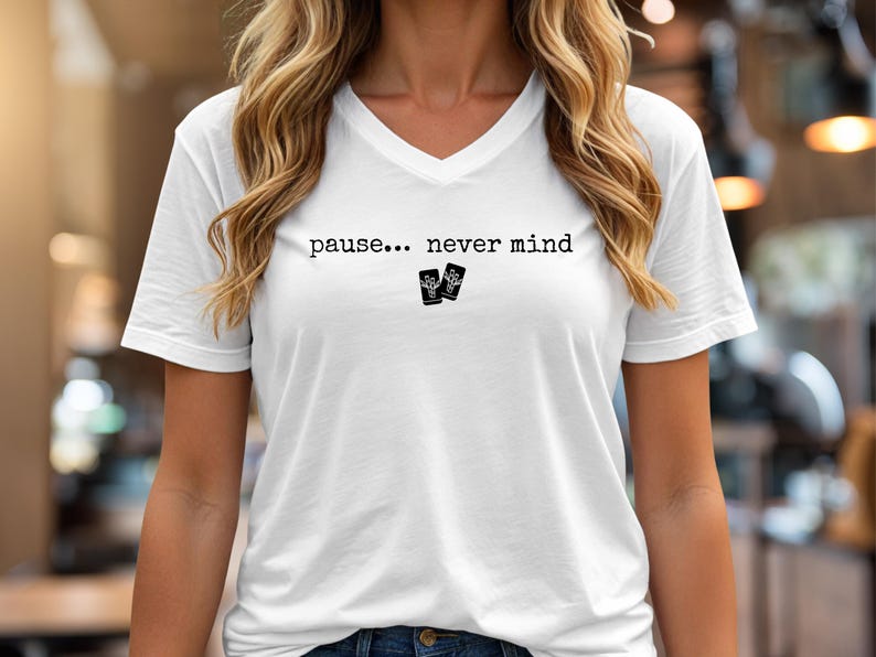 Pode incluir: Uma t-shirt branca com decote em V apresentando o texto preto "pause... never mind" impresso no peito. Abaixo do texto, h&aacute; dois pequenos &iacute;cones retangulares pretos, cada um exibindo um gr&aacute;fico branco de um cacto num vaso.