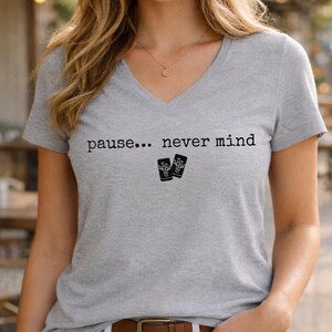 Pode incluir: Uma t-shirt cinzenta mesclada de manga curta com decote em V, apresentando o texto impresso a preto "pause... never mind" centrado no peito, acompanhado por um pequeno gr&aacute;fico de dois cart&otilde;es retangulares com uma m&atilde;o esquel&eacute;tica.
