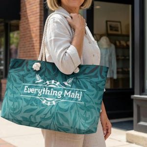 May include: A teal tote bag featuring a botanical leaf design and the white text "Everything Mahj". It has rope handles and white pom-pom details. The bag is being carried outdoors on a sunny day.