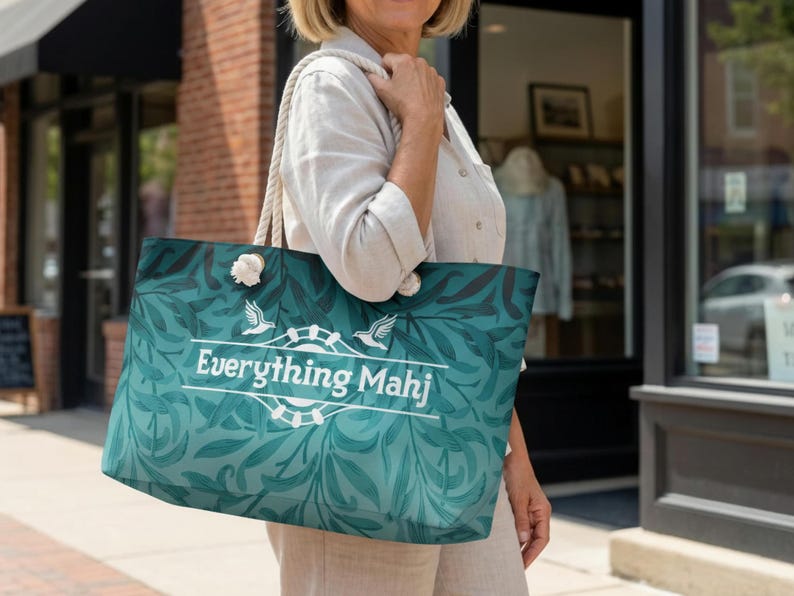 May include: A teal tote bag featuring a botanical leaf design and the white text "Everything Mahj". It has rope handles and white pom-pom details. The bag is being carried outdoors on a sunny day.